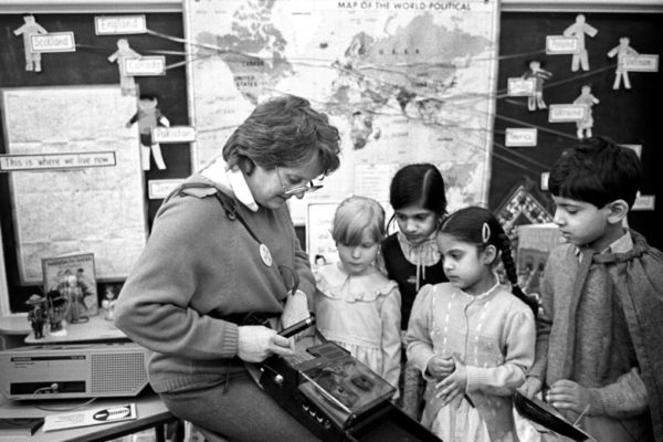 A black and white photograph depicting a teacher and pupils in a class room. They are standing in from of a world map. The teacher is holding a tap recorder while four children observe it.