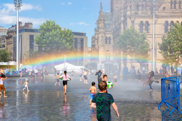 A rainbow over Bradford's City Park. Children are playing in the water fountain on a sunny day. In the centre of the photo is a large rainbow.