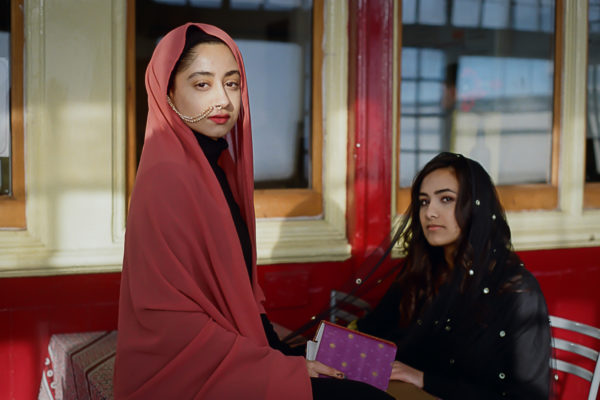 Two women called Sajidah and Mariyah from Belle Vue Girls Academy are posing for portraits. They have tried on some family heirlooms after interviewing their parents and grandparents about journeys from Pakistan to Bradford. They are seated in front of a bright red building. 